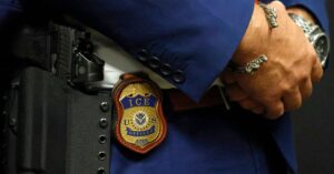 A photo shows a close-up of an Immigration and Customs Enforcement badge hanging from the waist of a federal agent. The agent, who is wearing a blue suit, a silver bracelet and a watch, holds their hands together.
