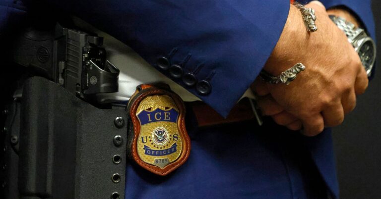 A photo shows a close-up of an Immigration and Customs Enforcement badge hanging from the waist of a federal agent. The agent, who is wearing a blue suit, a silver bracelet and a watch, holds their hands together.