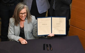 The image Gov. Katie Hobbs with shoulder-length gray hair and glasses, smiling while seated at a table. They are holding a blue-bordered document folder open, displaying two pages of text. The document is titled