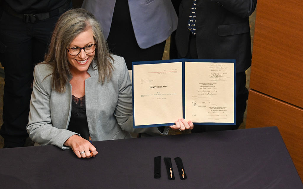 The image Gov. Katie Hobbs with shoulder-length gray hair and glasses, smiling while seated at a table. They are holding a blue-bordered document folder open, displaying two pages of text. The document is titled